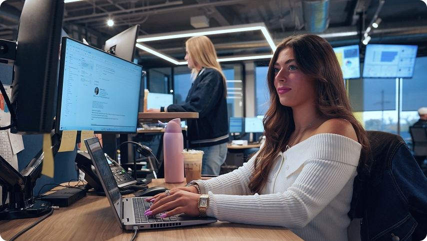 Female employee working on a laptop looking at monitors in an office